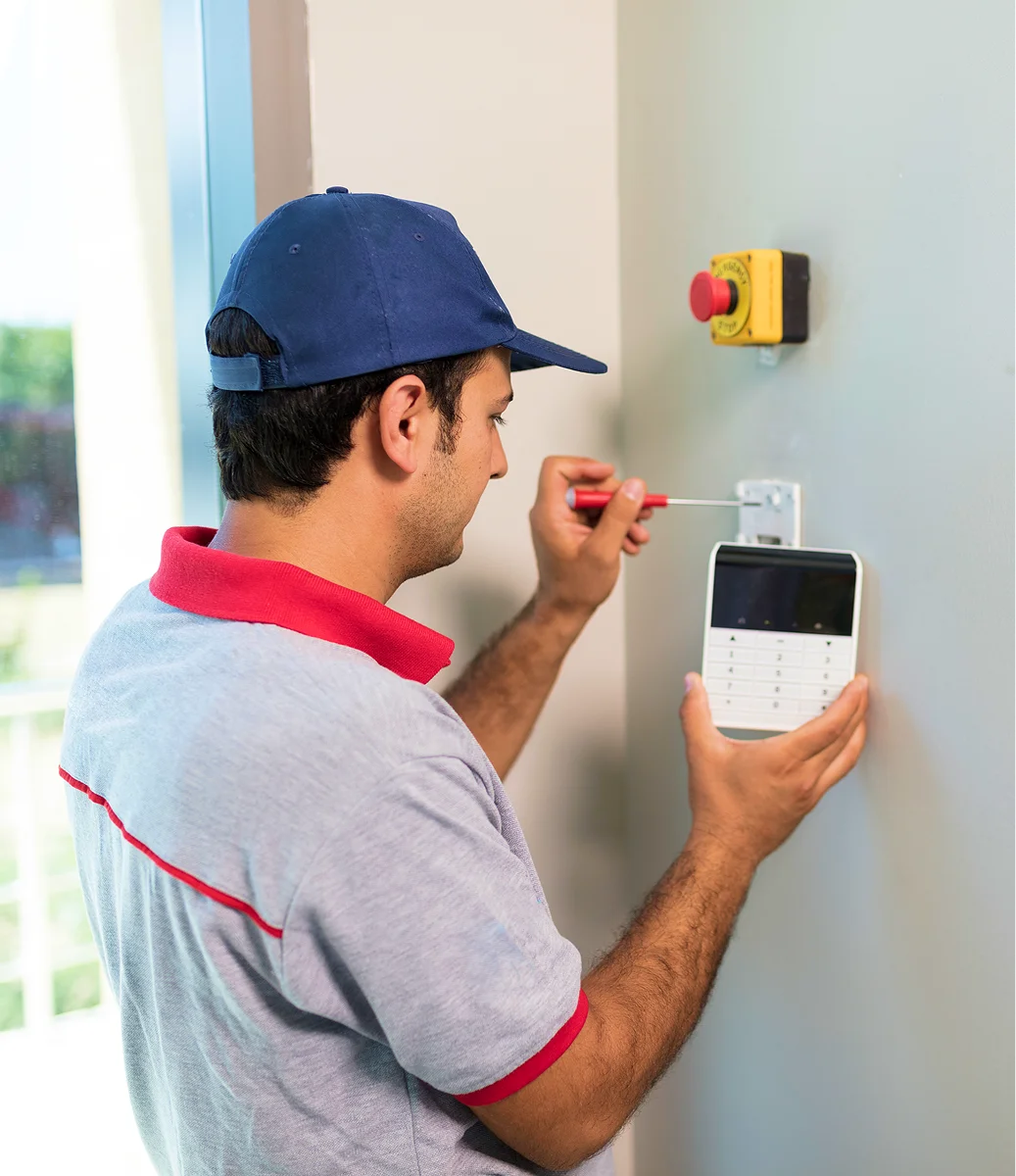 GettyImages-501047737 (1) Person wearing a blue cap and gray polo uses a screwdriver to install a security keypad on a wall beside an emergency button.