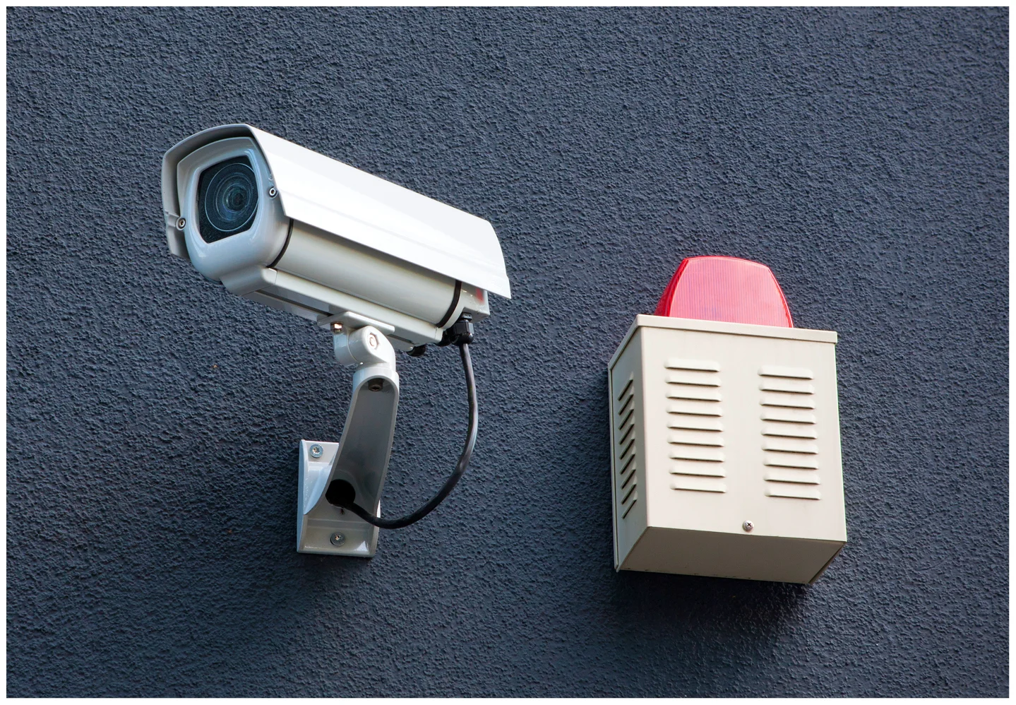 hero (35) A surveillance camera is mounted on a dark wall next to a beige alarm box with a red top.
